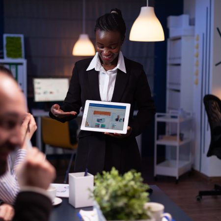 Entrepreneur woman with dark skin overworking in company office meeting room explaining management strategy using tablet. Workaholics multi-ethnic businesspeople brainstorming late at night.